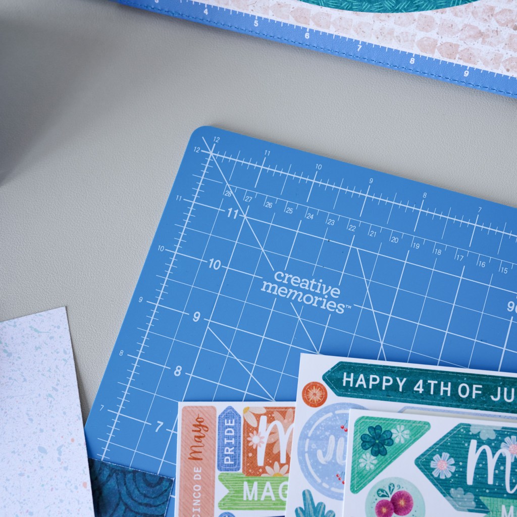 A creative workspace featuring a blue cutting mat with measurement lines, adorned with colorful decorative stickers for celebrations such as 'Happy 4th of July,' 'Mayo,' and 'Pride.'
