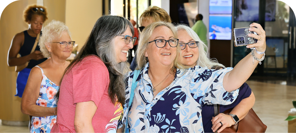 Three smiling women taking a selfie together in a bright indoor setting, with other people in the background.