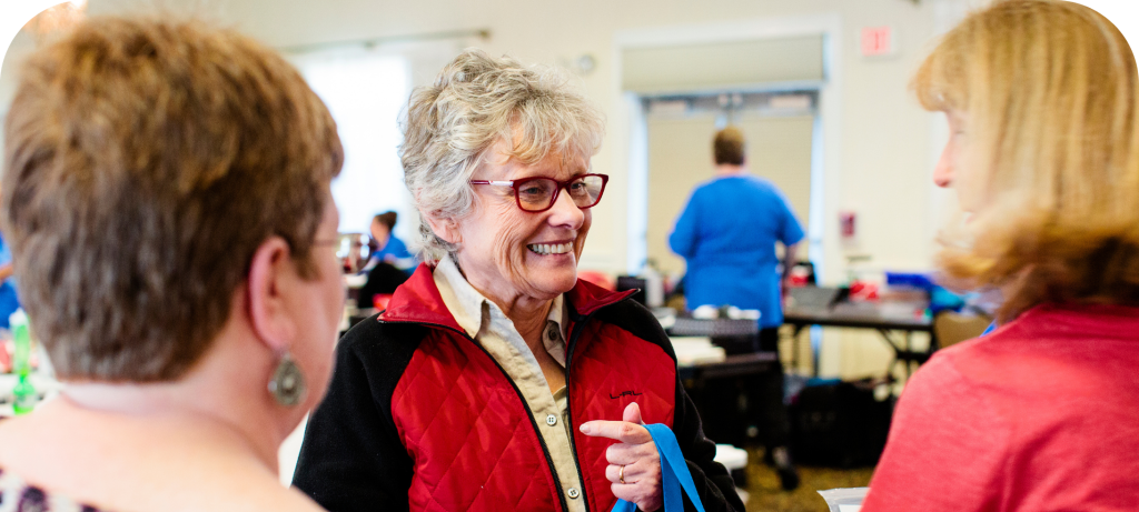Three women engaged in a conversation at a social event, with one woman laughing and smiling while pointing, indoors with other people in the background.