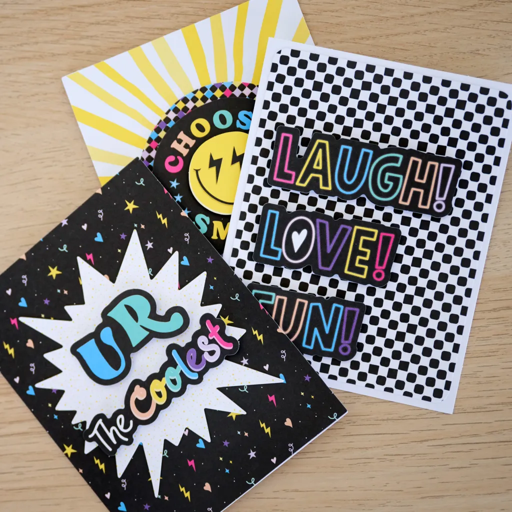 Three colorful greeting cards displayed on a wooden surface, featuring playful designs and phrases such as 'UR The Coolest,' 'LAUGH! LOVE! FUN!,' and 'Choose Smiles.'