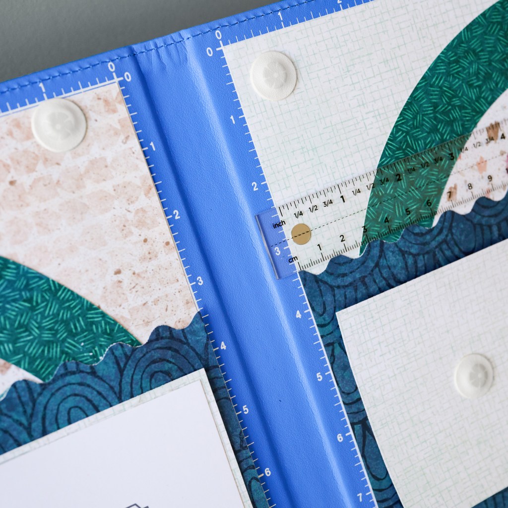 Close-up of colorful patterned papers arranged in a binder, featuring a ruler for measuring, with a blue background.