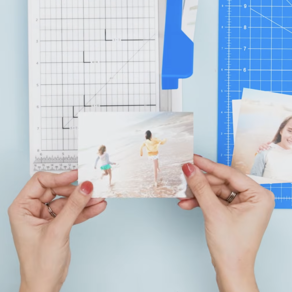A person holding a photograph of two children running on the beach, with crafting tools and a cutting mat in the background.