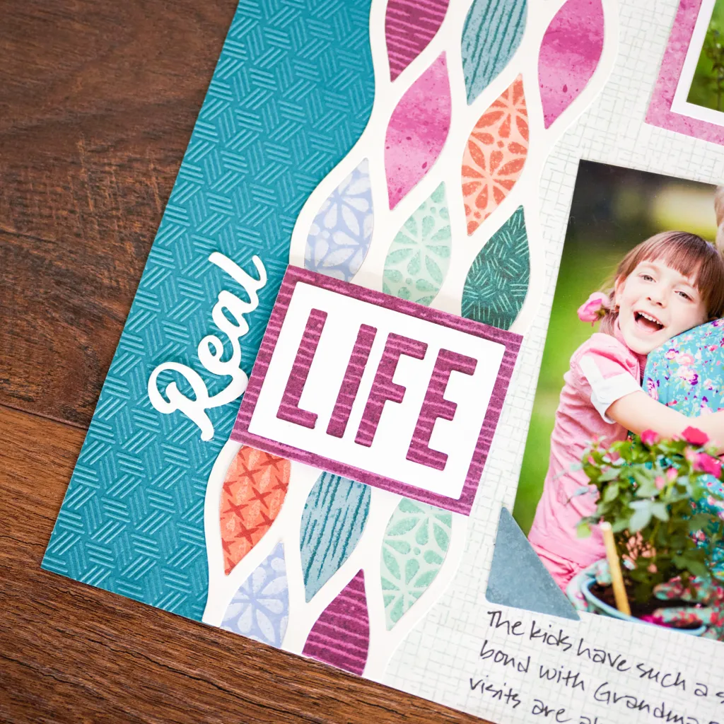 A close-up of a scrapbook page featuring the words 'Real LIFE' surrounded by colorful leaf patterns and a photo of a girl smiling.