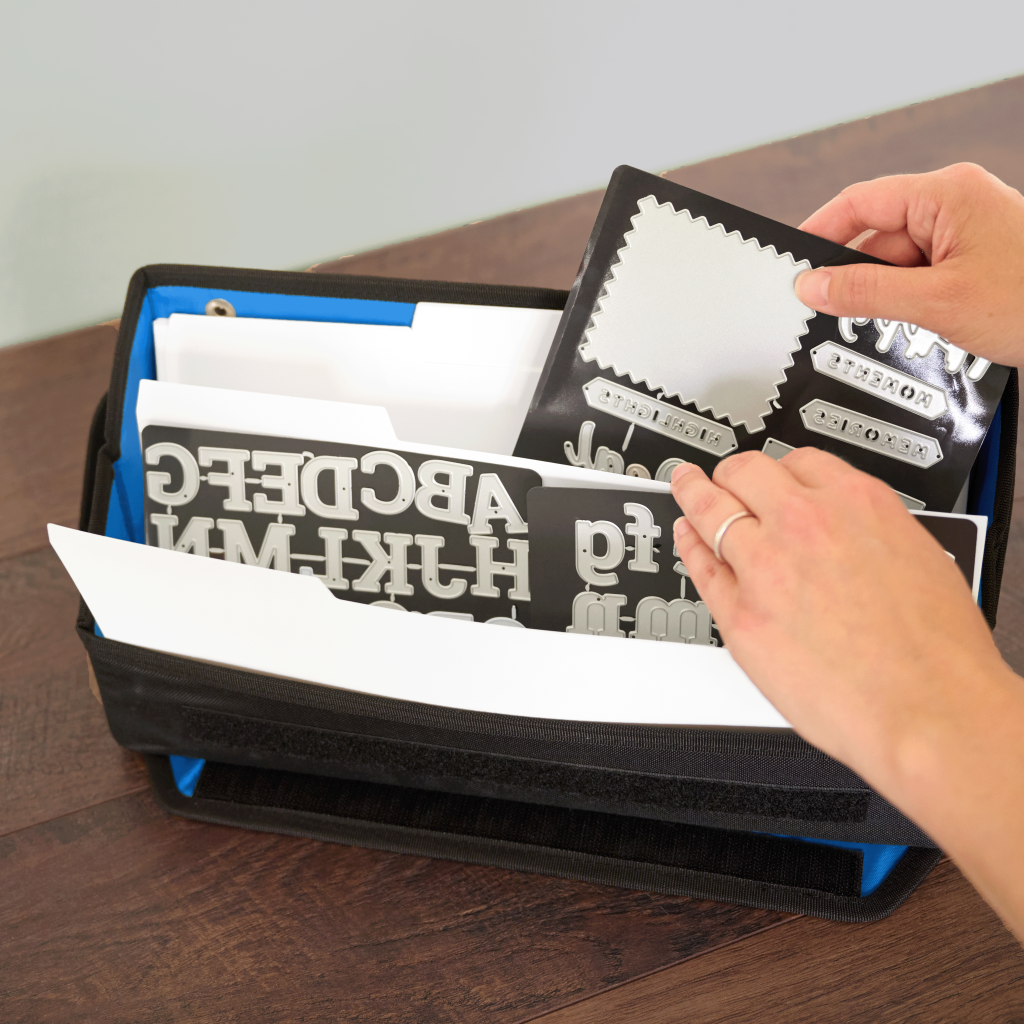 Person organizing crafting materials in a black storage box with blue lining, including paper and letter-shaped embellishments.
