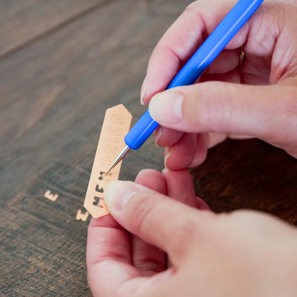 A person using a blue tool to engrave the word 'MEMORIES' on a copper tag placed on a wooden surface.