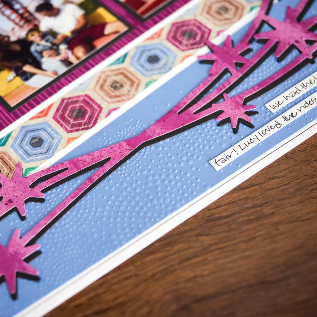 Close-up of a scrapbook page featuring colorful patterned paper, including hexagonal shapes and embossed blue texture, along with a pink starburst design and handwritten note.