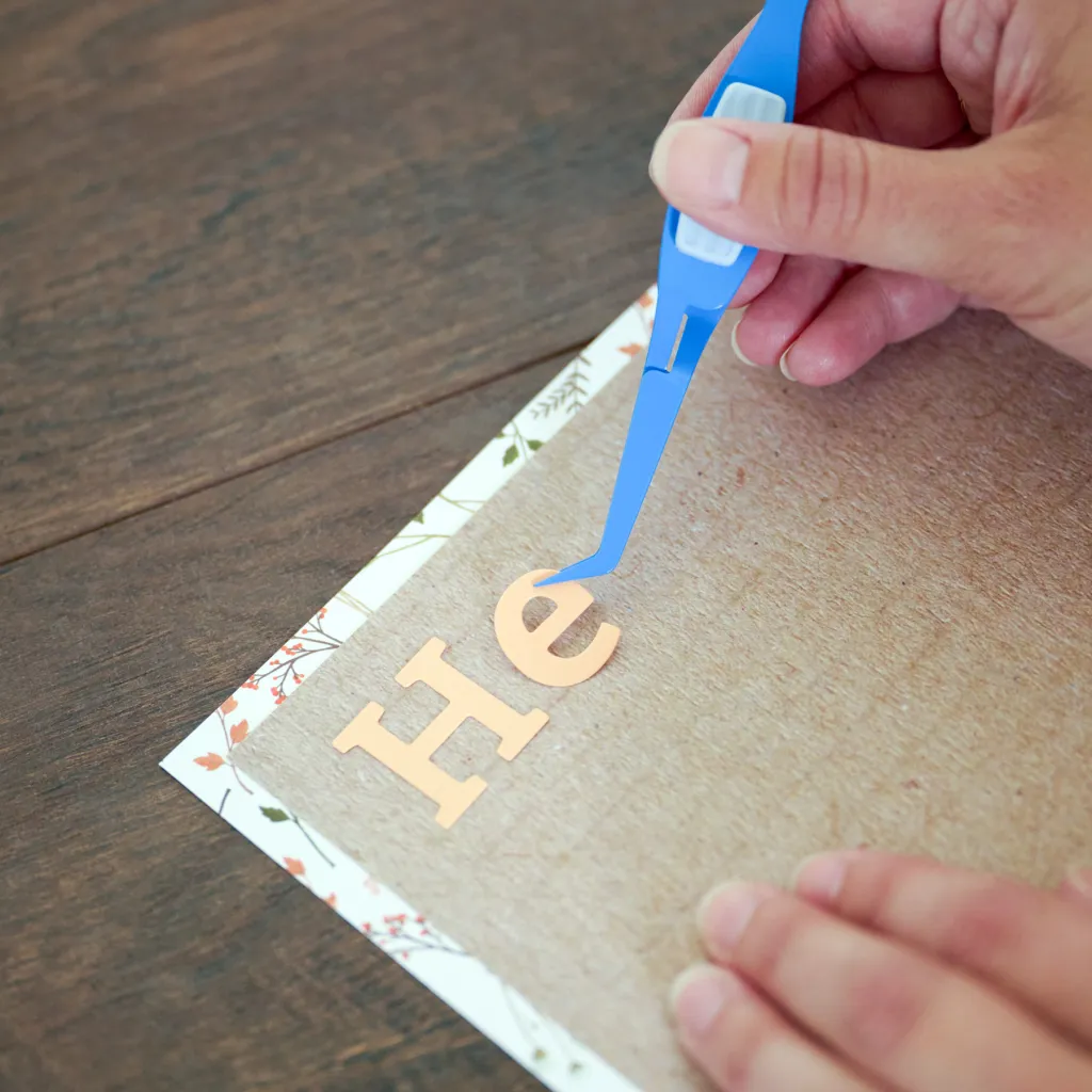 A person using a blue tweezer tool to place copper-colored letters onto a textured brown sheet of paper.