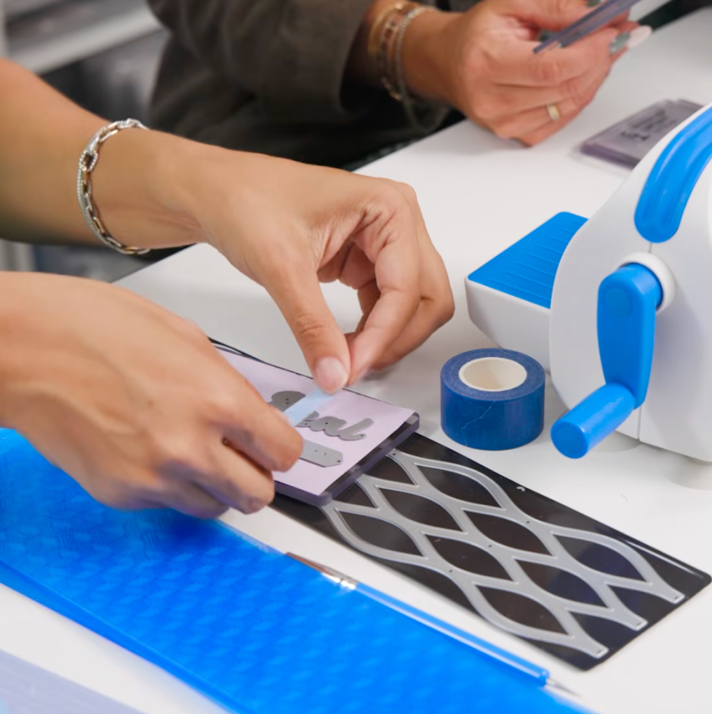 A person is using a crafting tool to position letters on a purple foam pad, surrounded by various crafting supplies including blue tape, a die-cut machine, and patterned sheets.